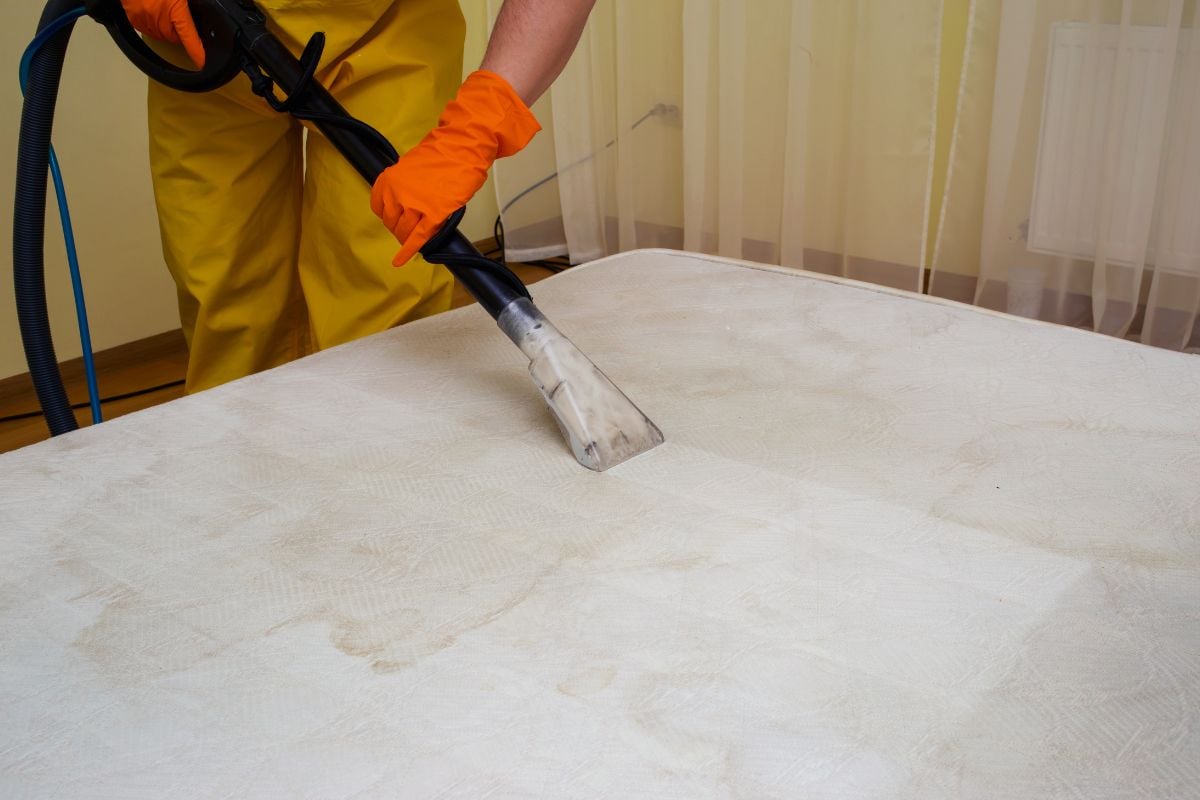 Person in yellow protective gear using a vacuum cleaner to clean a mattress, focusing on eliminating odors and maintaining a fresh sleep environment.