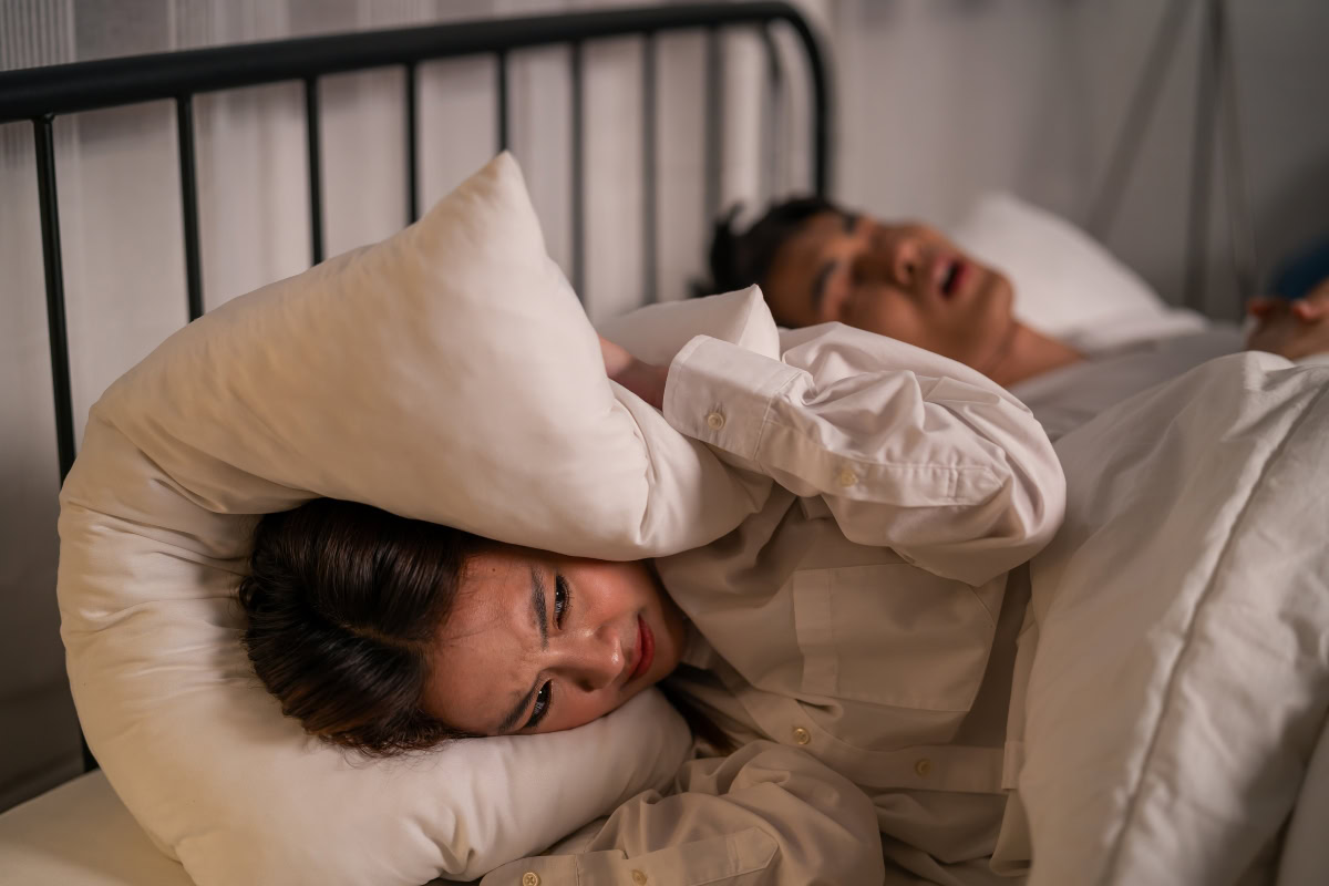 Person covering ears with pillow, distressed by partner's snoring in bed, illustrating challenges of sleep apnea and its impact on sleep quality.