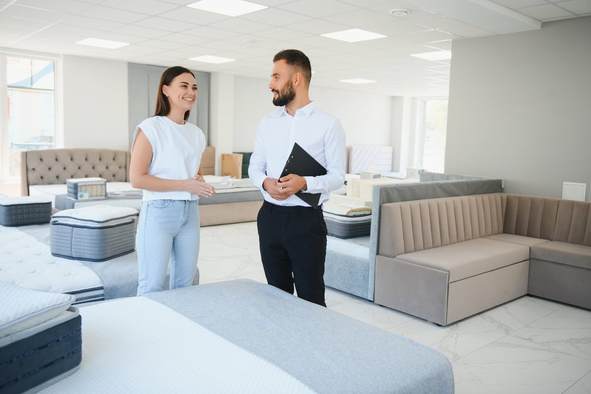 Smiling woman and man discussing affordable mattresses in a modern showroom with various mattress options displayed.