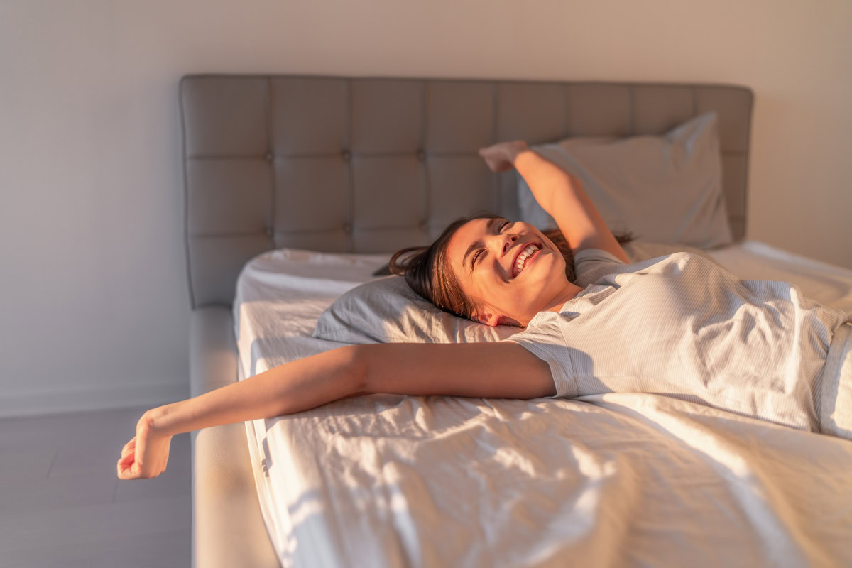 Woman smiling and stretching on an adjustable bed, showcasing comfort and relaxation in a well-lit bedroom setting.