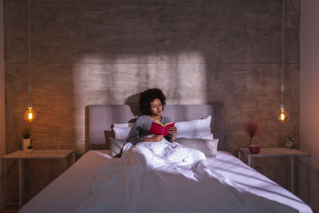 Cozy bedroom setting with a woman reading a book in bed, warm lighting, and minimalist decor, emphasizing a restful environment for better sleep.