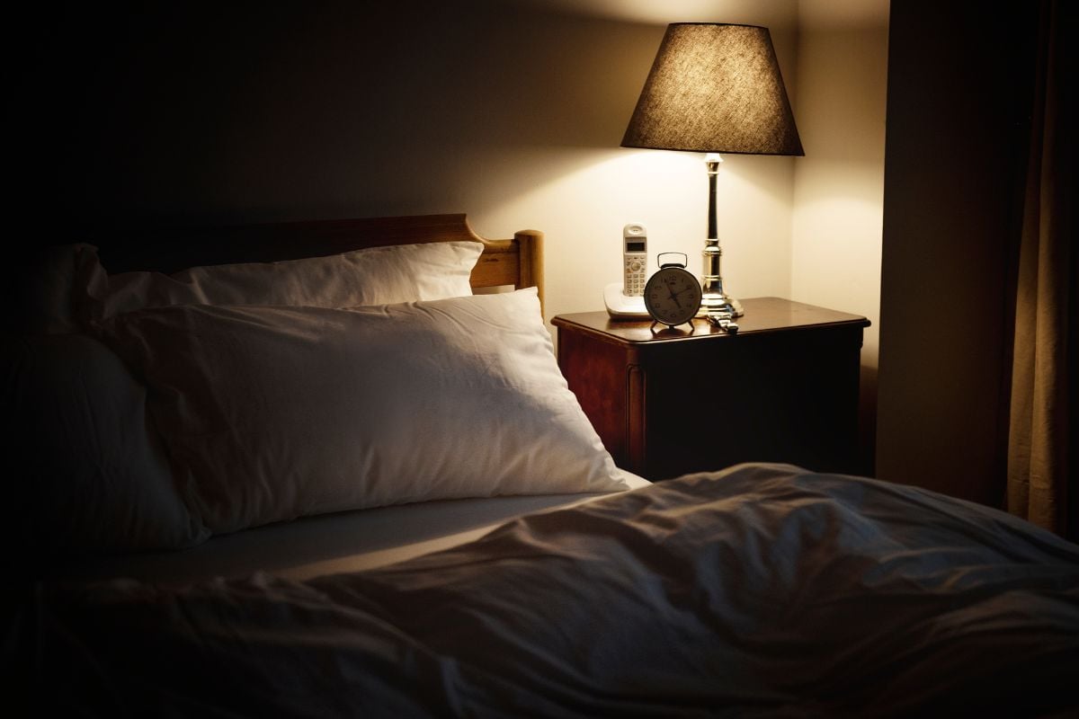 Cozy bedroom scene with soft lighting from a bedside lamp, neatly arranged pillows, and a clock on a nightstand, illustrating optimal room lighting for better sleep.